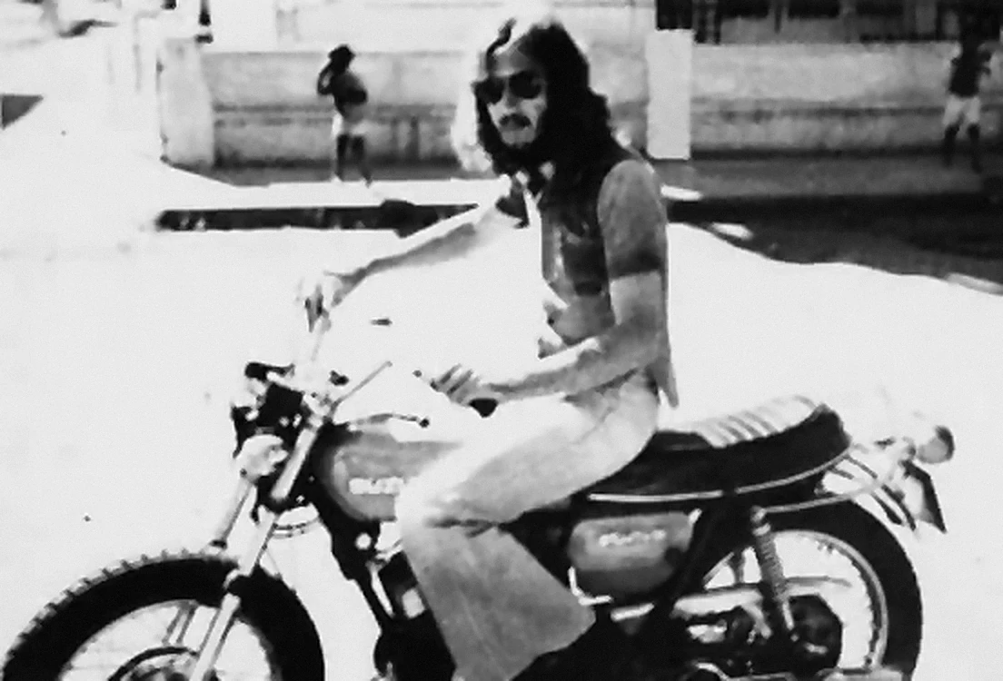 Fotografia antiga a preto e branco de um motociclista de cabelo comprido e óculos escuros, numa Suzuki